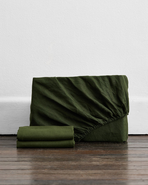 The Bed Threads Olive 100% French Flax Linen Fitted Sheet Set features a fitted sheet with visible elastic edging and two folded flat sheets, shown on a wooden floor—essential bedding for any room.