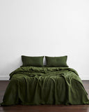 A neatly made bed with dark green bedding and two Bed Threads Olive 100% French Flax Linen Pillowcases, set against a plain white wall and a dark wooden floor.