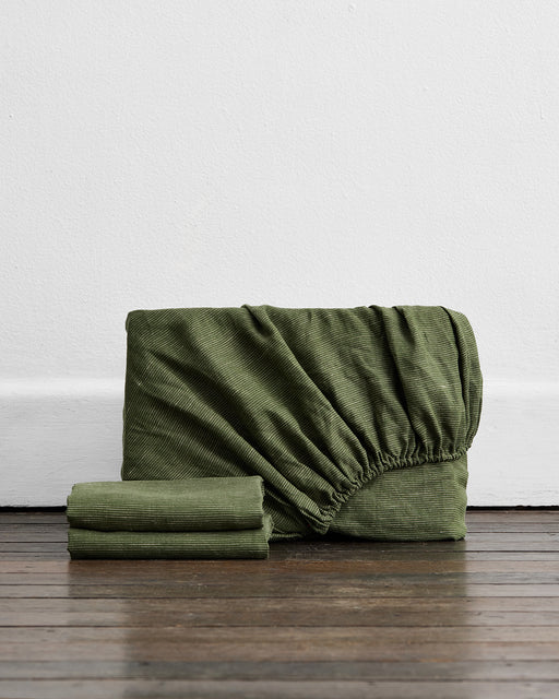 The Bed Threads Olive Stripe 100% French Flax Linen Fitted Sheet Set, including pillowcases, is neatly folded on a wooden floor against a plain white wall, adding cozy charm to any linen bedding collection.