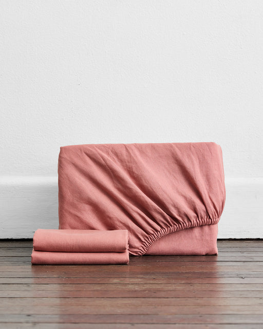 The Bed Threads Pink Clay 100% French Flax Linen Fitted Sheet Set, featuring a folded fitted sheet with elastic edges and two matching pillowcases, is displayed upright on a wooden floor against a white wall.
