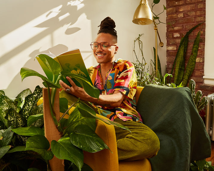 A person wearing glasses and a colorful shirt sits in a yellow chair surrounded by lush green plants, smiling while reading a book in a sunlit room.