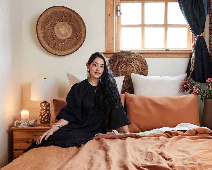 A woman with long dark hair in a black dress sits on a bed with brown and white bedding in a cozy, well-decorated bedroom. Woven baskets hang on the wall and a lit candle sits on a wooden nightstand.