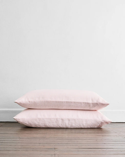 Two Rosewater 100% French Flax Linen King Pillowcases by Bed Threads are stacked against a white wall on a wooden floor, adding a soft pink touch to this minimal, bright room.