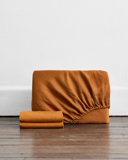 A Rust 100% French Flax Linen Fitted Sheet Set by Bed Threads, featuring a fitted sheet with visible elastic edge and two matching pillowcases, is neatly folded on a wooden floor against a white wall.