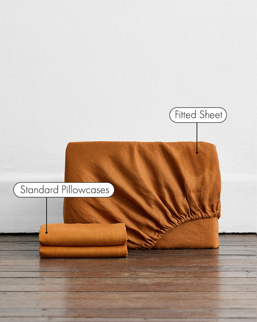 A Rust 100% French Flax Linen Fitted Sheet Set by Bed Threads is folded upright on a wooden floor against a white wall, with two matching standard pillowcases nearby. Labels highlight the fitted sheet and pillowcases.