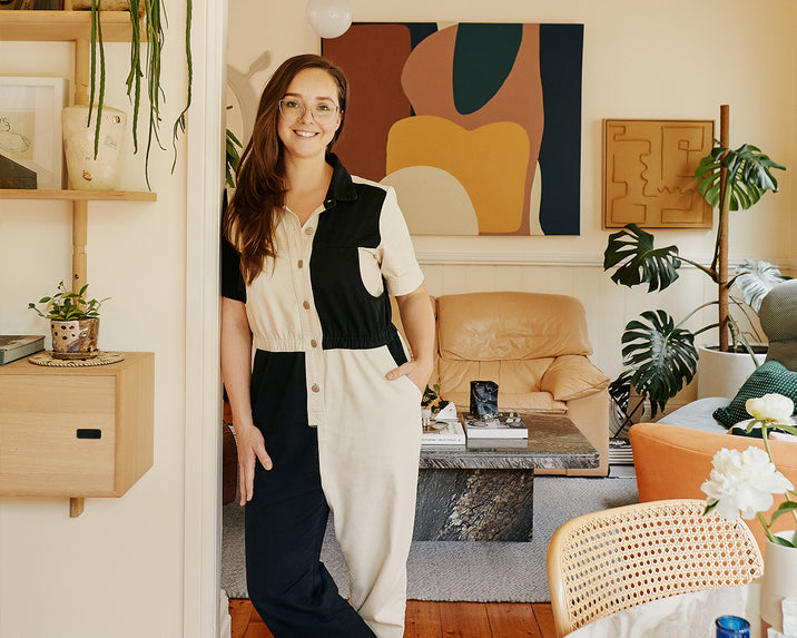 A woman in a black and white jumpsuit stands smiling in a cozy, art-filled living room with plants, a tan couch, and abstract wall art.