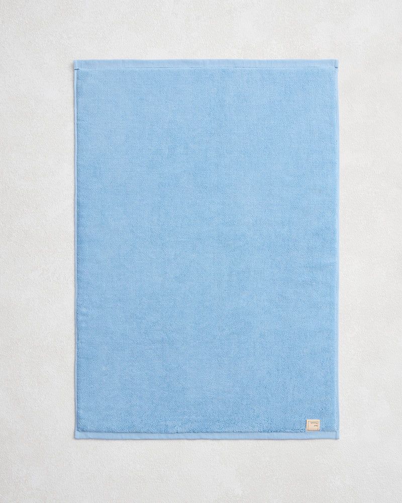 The Bed Threads Sky 100% Cotton Terry Bath Mat, in light blue, lies flat and neatly folded on a pale textured surface with a small fabric label attached to one corner.