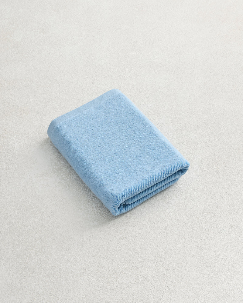 A Sky 100% Cotton Terry Bath Mat by Bed Threads rests on a light, textured surface beside a neatly folded blue towel, bringing comfort and style to your bathroom.