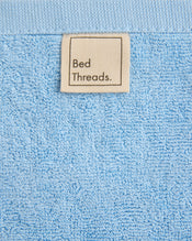 A close-up of the Sky 100% Cotton Terry Bath Mat by Bed Threads shows its soft, textured surface and a beige fabric label with "Bed Threads." in black text. The mat's plush texture highlights its luxurious quality. - view