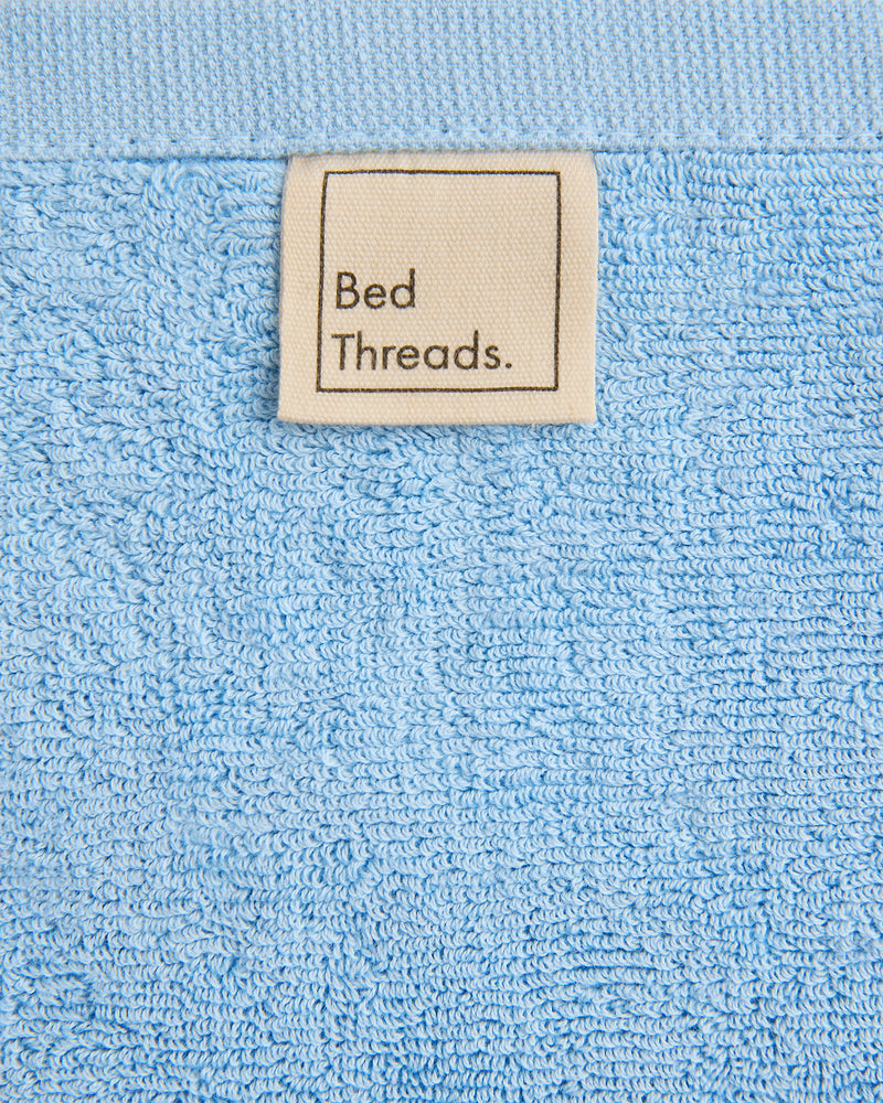 A close-up of the Sky 100% Cotton Terry Bath Mat by Bed Threads shows its soft, textured surface and a beige fabric label with 