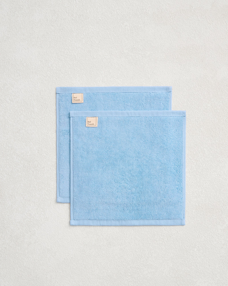 A set of two Sky 100% Cotton Terry Face Washers by Bed Threads, in light blue with small beige labels, are slightly overlapping on a textured surface—ideal for your daily skincare routine.
