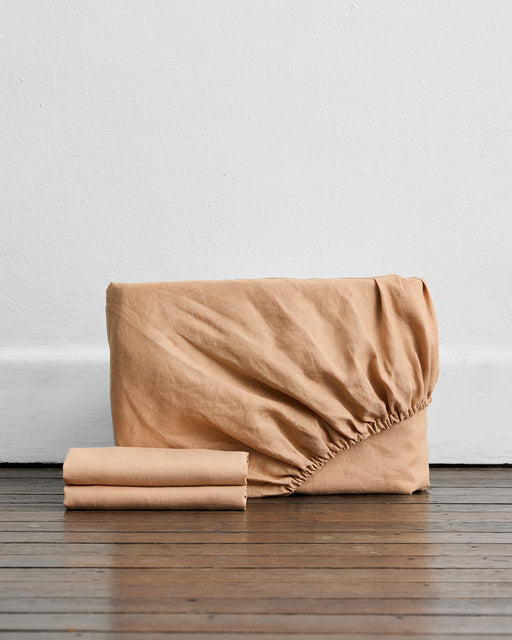 A stack of Bed Threads' Terracotta 100% French Flax Linen Fitted Sheet Set, featuring a partially unfolded fitted sheet and two neatly folded flat sheets, sits on a dark wooden floor against a white wall—true bedding essentials.