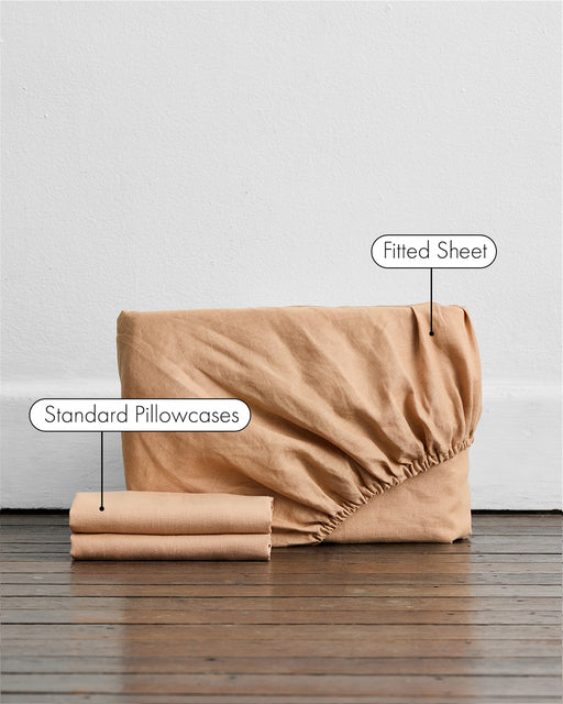 A Terracotta 100% French Flax Linen Fitted Sheet Set by Bed Threads, including a folded fitted sheet and two standard pillowcases, rests against a wall on a wooden floor. Labels indicate the Fitted Sheet and Standard Pillowcases.