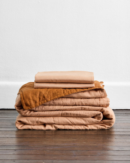 A neatly folded stack of the Bed Threads Terracotta & Rust Quilt Set sits on a wooden floor against a white wall.