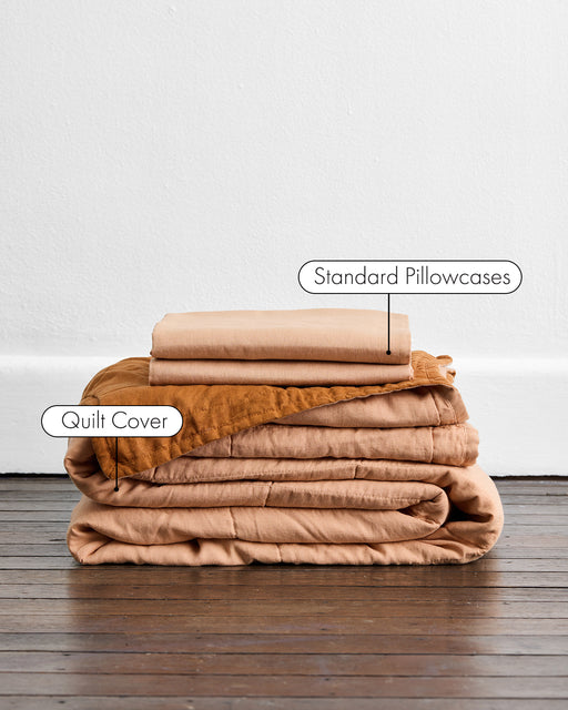 A stack of Bed Threads’ Terracotta & Rust Quilt Set sits on a wooden floor against a white wall, with standard pillowcases on top and a quilted bed cover labeled Quilt Cover below.