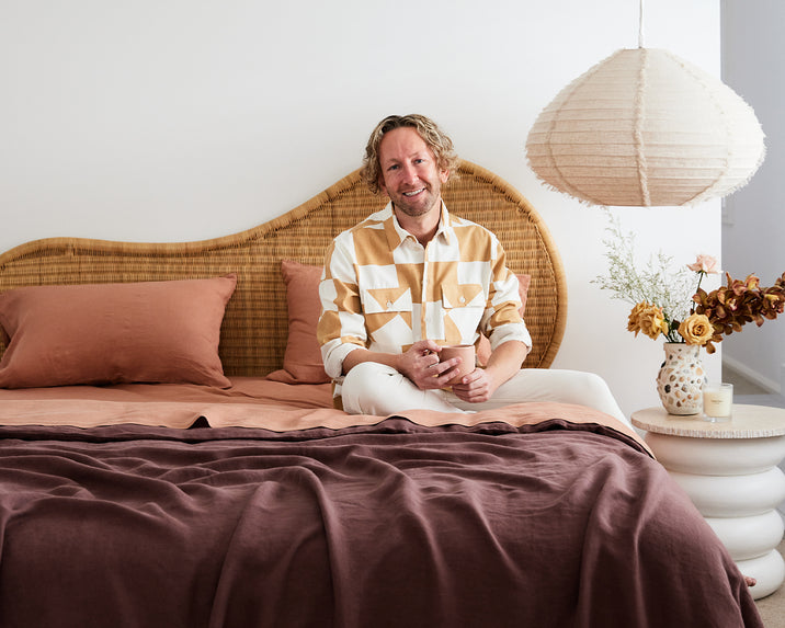 A smiling man with wavy blonde hair sits on a made bed with rust and mauve bedding, against a rattan headboard, holding a mug. A paper lantern and a vase with flowers are on a bedside table beside him.