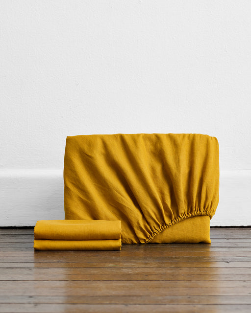 A Turmeric 100% French Flax Linen Fitted Sheet and two folded pillowcases from Bed Threads’ Fitted Sheet Set are displayed on a wooden floor against a plain white wall.