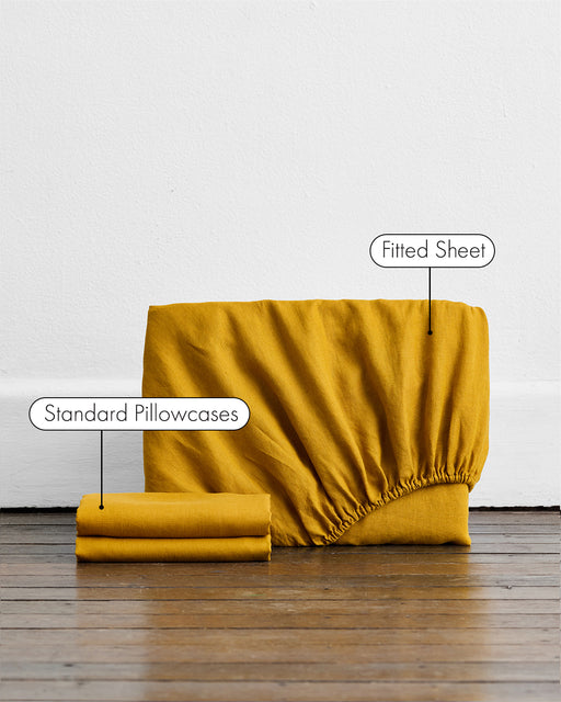 A folded Bed Threads Turmeric 100% French Flax Linen Fitted Sheet Set, with two matching pillowcases, is labeled and displayed on a wooden floor against a white wall.