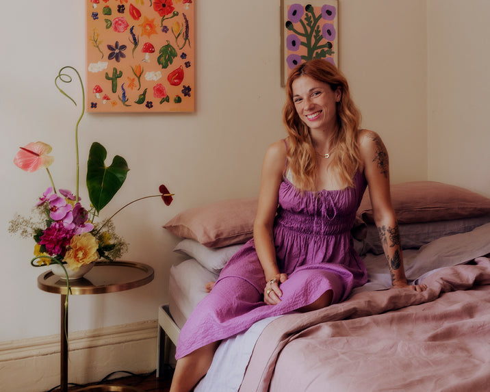 A woman with long, wavy hair in a purple dress sits on a bed with pink sheets, smiling. Behind her are colorful floral paintings and a small round table with a vibrant flower arrangement.