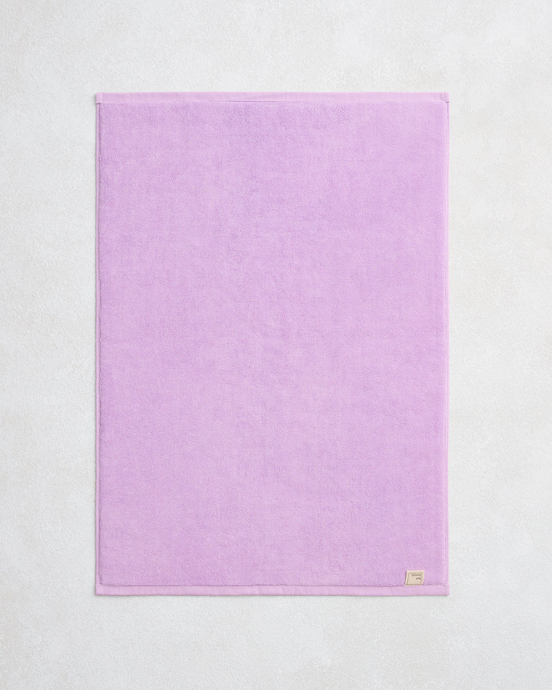 The Bed Threads Violet 100% Cotton Terry Bath Mat, featuring a small fabric tag in the bottom right corner, is neatly arranged and lies flat on a light textured surface.