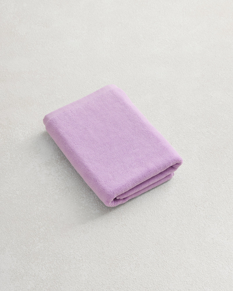 The Bed Threads Violet 100% Cotton Terry Bath Mat, in a soft lavender shade, sits on a light textured surface next to a neatly folded matching towel for ultimate comfort and luxury.
