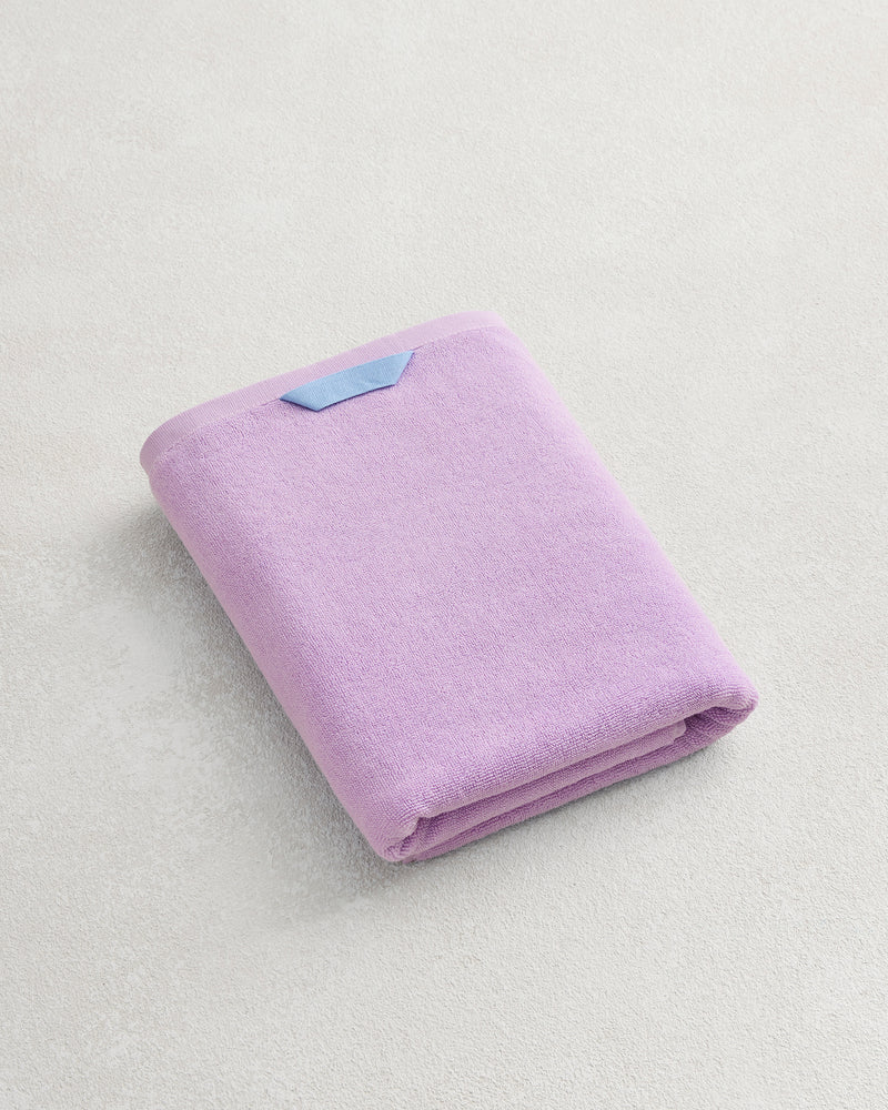 The Violet 100% Cotton Terry Bath Towel by Bed Threads, featuring a blue tag, rests on a light textured surface—ideal for those who want quick-drying, highly absorbent towels.