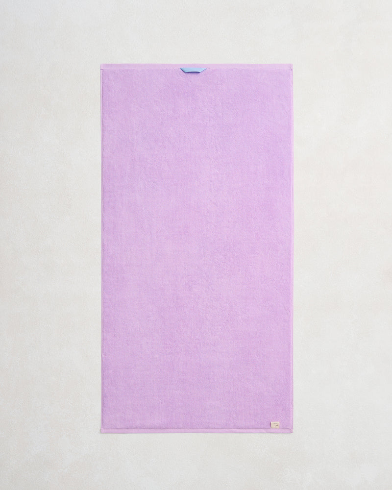 The Bed Threads Violet 100% Cotton Terry Bath Towel is a neatly rectangular, light purple towel with a blue loop on the top edge and a beige tag at the bottom right—ideal for those seeking absorbent, stylish bath linens.