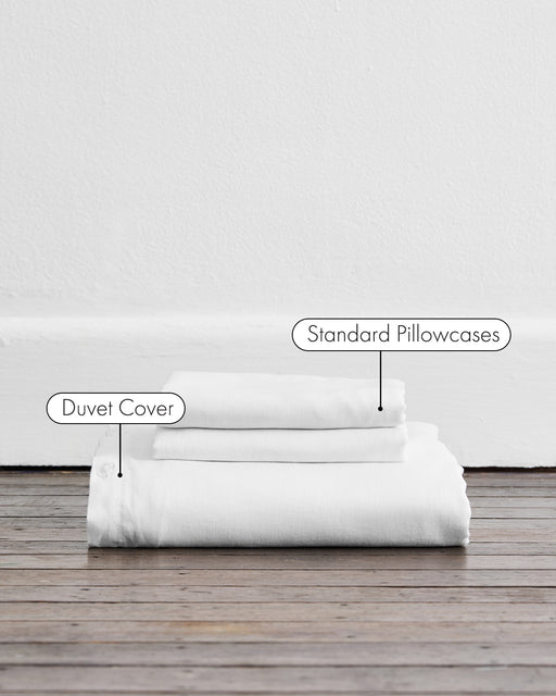 A folded White 100% French Flax Linen Duvet Cover Set by Bed Threads, with matching pillowcases, is stacked on a wooden floor against a white wall—ideal for anyone seeking luxurious bedding essentials.