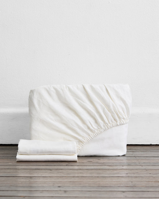 A folded White 100% French Flax Linen Fitted Sheet with gathered elastic stands upright beside two pillowcases from the Bed Threads Fitted Sheet Set, all set on a wooden floor against a plain white wall.
