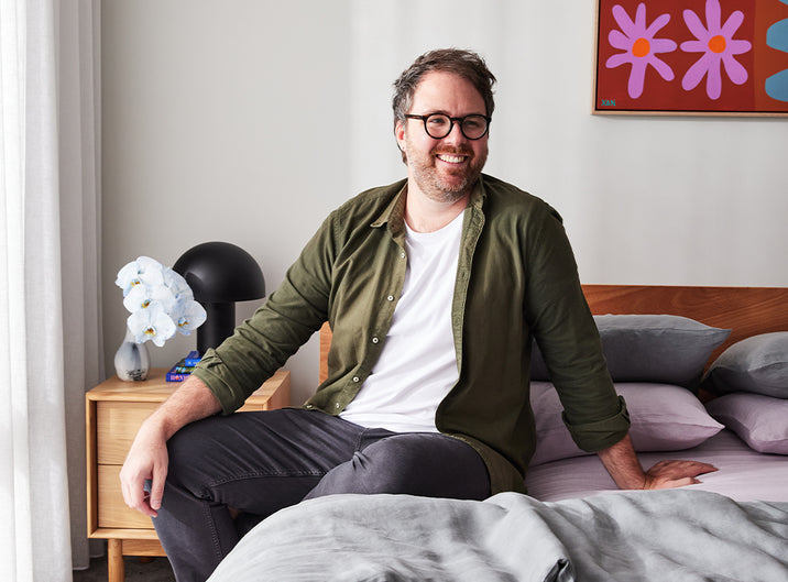 A smiling man with glasses sits on a bed in a modern bedroom with gray bedding, a bedside table, a black lamp, white orchids, and colorful floral artwork on the wall.