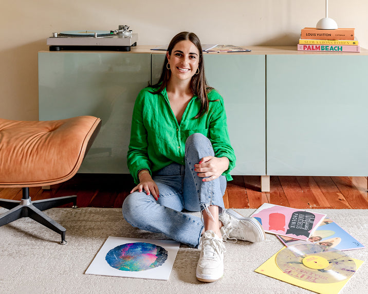 A woman in a bright green shirt and jeans sits cross-legged on a rug, smiling. She’s surrounded by colorful vinyl records, next to a tan chair, with a turntable and stacked books on a cabinet behind her.