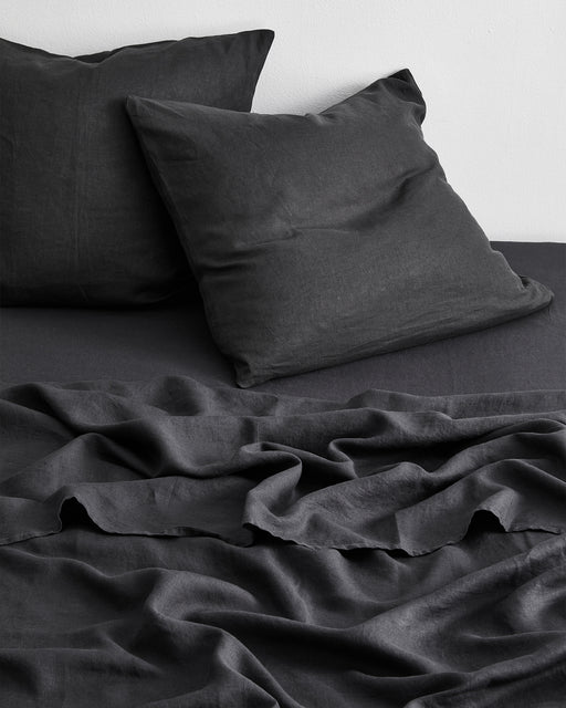 A neatly made bed features two Bed Threads Charcoal 100% French Flax Linen European Pillowcases, paired with a matching dark gray sheet and blanket, all slightly wrinkled against a plain white wall.