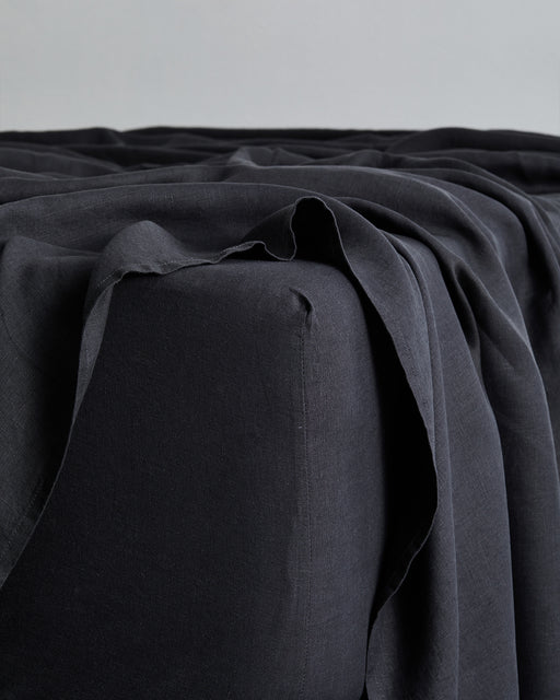 A close-up of a dark piece of furniture draped with the Bed Threads Charcoal 100% French Flax Linen Fitted Sheet, featuring soft folds and a slightly wrinkled, minimal modern look.