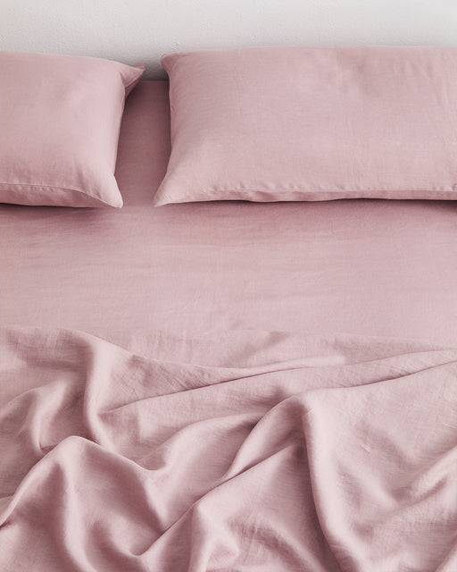A neatly made bed features two pillows and a slightly wrinkled Bed Threads Lavender 100% French Flax Linen Fitted Sheet in soft pink. The bedding looks clean, cozy, and offers temperature regulation for enhanced comfort.