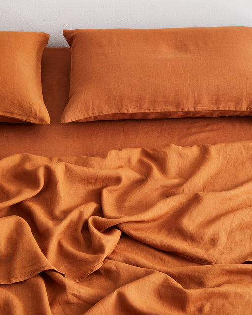 A neatly made bed features two pillows and a Bed Threads Rust 100% French Flax Linen Fitted Sheet in a warm, rust-orange hue; the rumpled fitted sheet adds inviting texture to the scene.