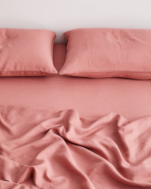 A bed features two pink pillows and a Pink Clay 100% French Flax Linen Fitted Sheet by Bed Threads, its slightly rumpled surface creating soft, temperature-regulating folds at the foot.