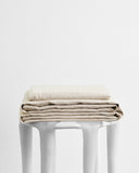 The Bed Threads Oatmeal 100% French Flax Linen Sheet Set is folded atop a sleek white stool against a plain backdrop, highlighting its temperature-regulating benefits in an elegant, minimalist display.