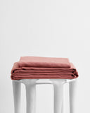 A neatly folded stack of Bed Threads Pink Clay 100% French Flax Linen Sheet Set rests on a modern white stool set against a plain white background.