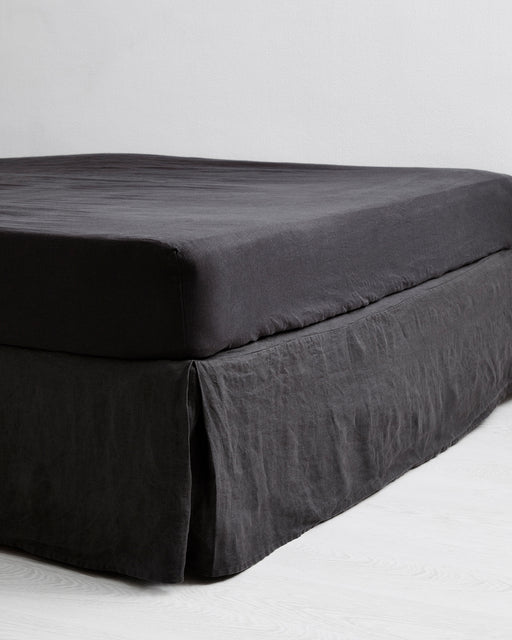 A neatly made bed features a dark gray blanket, two matching pillows, and a Bed Threads Charcoal 100% French Flax Linen Valance, all set against a plain white wall on a light-colored floor.