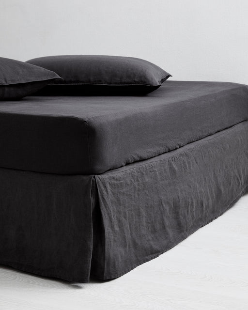 A neatly made bed with dark gray bedding, including a pillow, fitted sheet, and the Bed Threads Charcoal 100% French Flax Linen Valance, is set against a plain white wall on a light-colored floor.