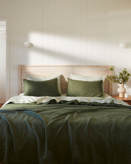 A neatly made bed featuring the Bed Threads Olive & Sage 100% French Flax Linen Bedding Bundle, accented by green and cream pillows, a wooden headboard, a bedside table with greenery, and white paneled walls in a minimalist bedroom.