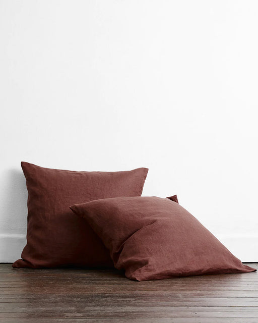 Two square brown pillows in Bed Threads' Cacao 100% French Flax Linen European Pillowcases (Set of Two) rest on a dark wooden floor against a white wall—one upright, the other gently leaning in front.