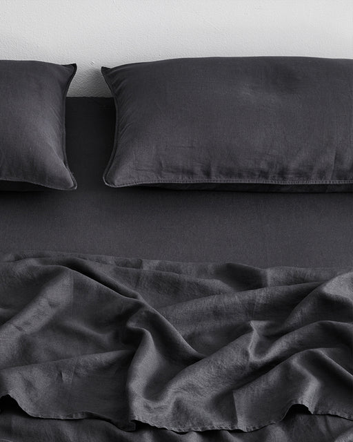 A neatly made bed with two dark gray pillows and a slightly wrinkled Bed Threads Charcoal 100% French Flax Linen Fitted Sheet rests against a light wall.