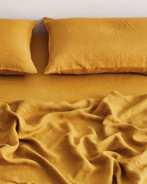 Two mustard yellow pillows and a Bed Threads Turmeric 100% French Flax Linen Fitted Sheet rest on a bed, viewed from above. The textured linens add warmth and coziness, creating an inviting atmosphere perfect for relaxing.