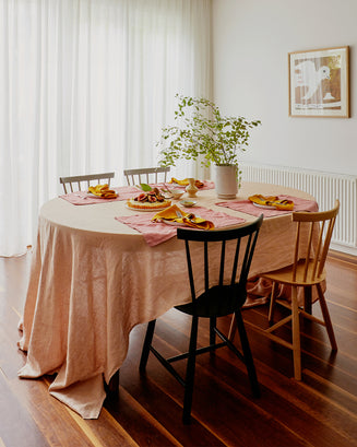 A round dining table is set for four with the Bed Threads Terracotta 100% French Flax Linen Tablecloth, pink napkins, plates of food, and mismatched wooden chairs. A leafy plant centerpiece, sheer curtains, and art finish the look.